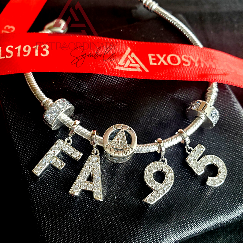 Product photography of a silver charm bracelet featuring encrusted F A 9 5 letter charms and a round "est 1913" symbol charm, resting on black fabric with a red EXOSYMBOLS ribbon